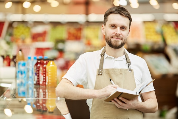 Waist up portrait of bearded man wearing apron and holding notebook while standing in supermarket