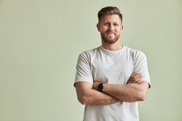 Waist up portrait of bearded young man smiling at camera while standing with arms crossed against pa