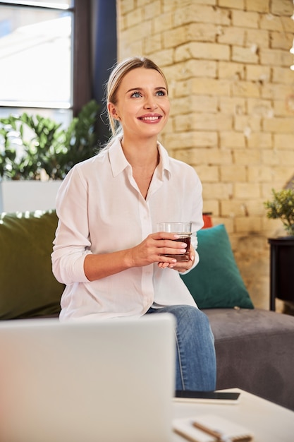 Waist up portrait of charming business lady holding glass with drink in hands while enjoying good time in the beautiful meeting room