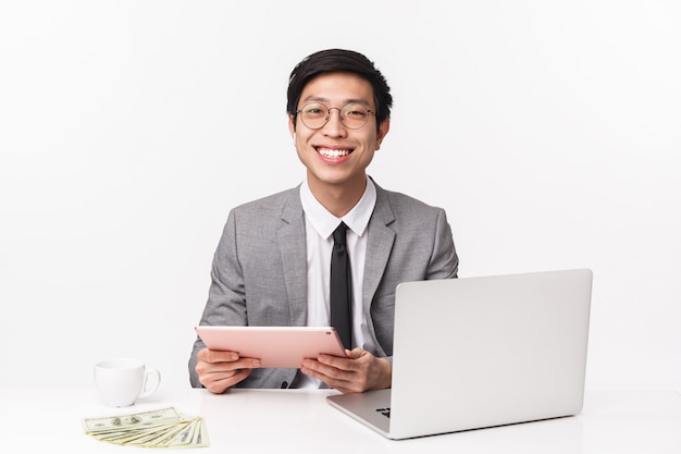 Талия-вверх портрет веселого, счастливого улыбающегося азиатского офисного работника в костюме, сидя за столом, изучая диаграмму с помощью цифрового планшета и ноутбука, отвечая на звонки с беспроводными наушниками, на белой стене