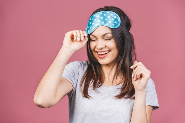 Waist up portrait of cheerful smiling girl in sleeping mask. Attractive funny female in stylish pajama standing and looking away. Isolated on pink background.