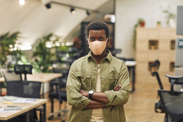 Waist up portrait of confident African-American man wearing mask in office and  while standing with arms crossed in office, copy space