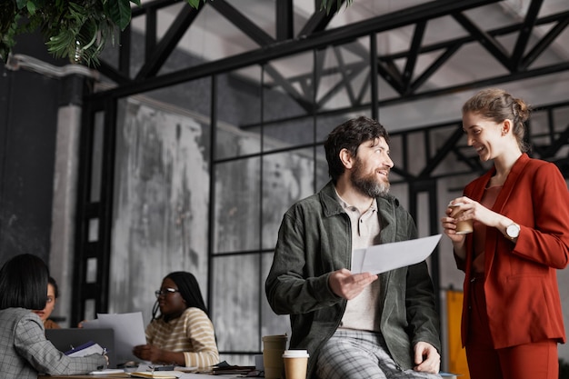 Waist up portrait of contemporary bearded businessman talking to female colleague and holding documents in graphic office interior, copy space