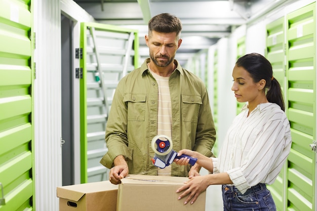 Waist up portrait of contemporary couple packing boxes with tape gun while standing in self storage unit, copy space