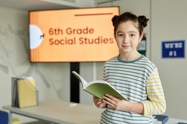 Waist up portrait of cute schoolgirl in striped shirt looking at camera and holding book while stand