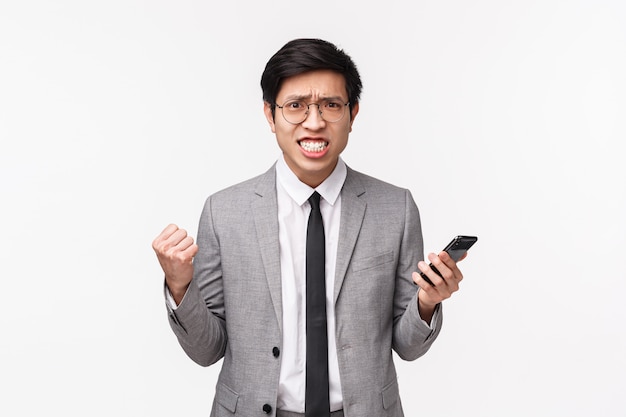 Waist-up portrait of displeased, bothered and annoyed asian businessman in grey suit, clenching teeth and fists, frowning aggressive, grimacing irritated from reading bad news on mobile phone