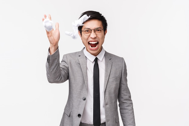 Waist-up portrait of distressed, stressed and angry young asian male manager, office worker destroy his report, decide to quit, tear apart documents and throw them in air, shouting outraged