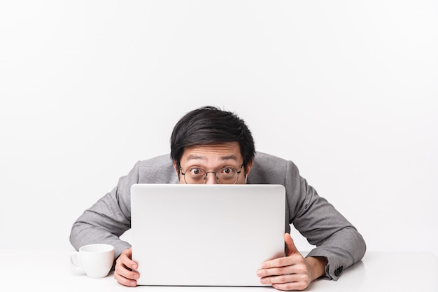 Waist-up portrait of funny and silly handsome asian office worker, man in suit sitting at table hiding face behind opened laptop, peeking at , avoiding work and fool around, on a white wall