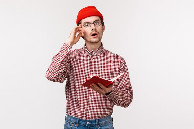 Waist-up portrait geeky and creative guy in red beanie, glasses have stunning great idea, touch temple and look inspired while taking notes in notebook, creating equation, stand on a white wall