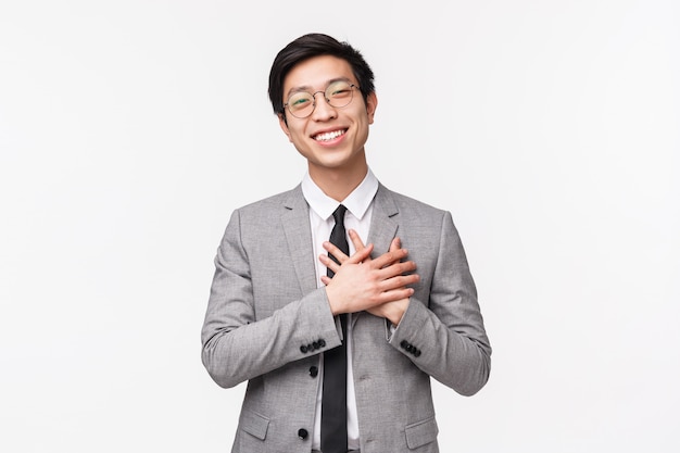 Waist-up portrait of grateful, flattered asian young male office manager, entrepreneur hold hands on heart and smiling, thanking for compliment, flattered and grateful for praises, on a white wall