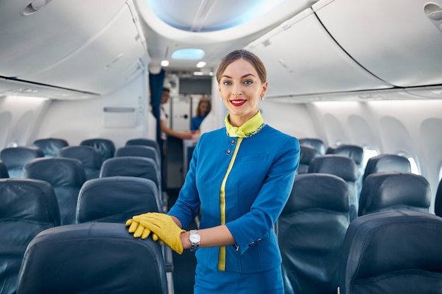 Waist up portrait of happy cheerful female wearing flight attendant blue uniform with yellow leather gloves posing at the camera on the empty board