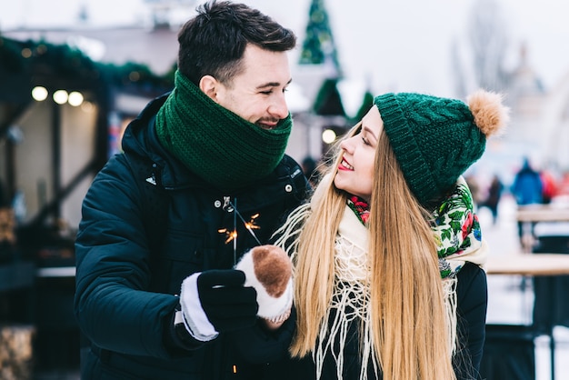 Waist up portrait of happy loving couple admiring winter weather in park. They are cuddling and laughing while looking at each other. People holding sparkles