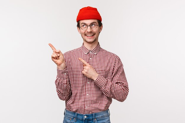 Waist-up portrait of happy smiling geeky guy in glasses and red beanie, pointing and looking upper left corner with pleased grin, satisfied found good product, recommend use this service