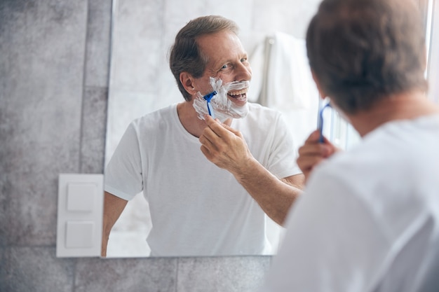 Waist-up portrait of a high-spirited man with a short haircut shaving himself with a razor