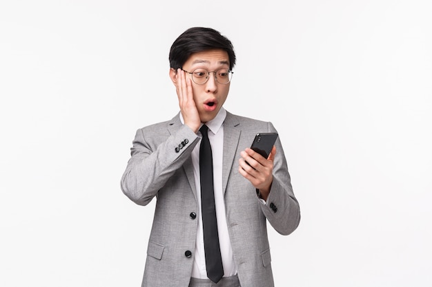 Waist-up portrait of impressed, speechless asian handsome businessman in grey suit, gasping touch cheek amazed as staring at smartphone display, reading shocking news or message