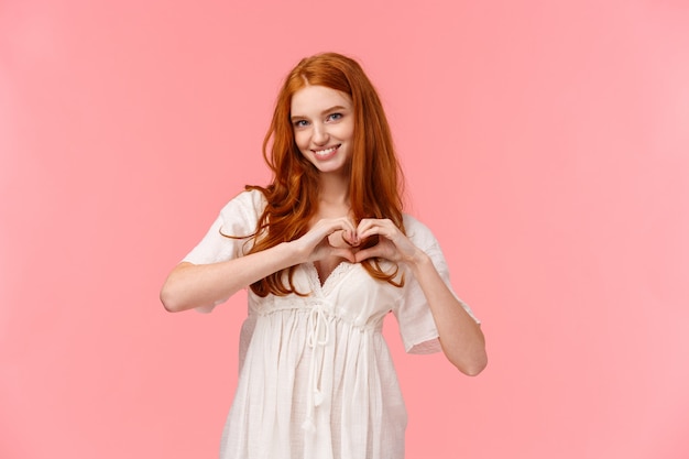 Waist-up portrait lovely, sensual redhead girlfriend in white dress, showing her love and care, make heart gesture and smiling, confess affection, sincere sympathy, pink wall