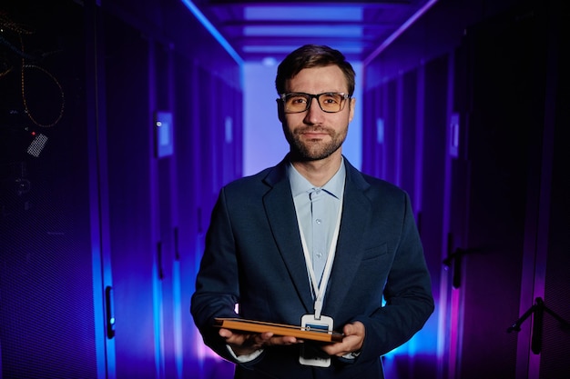 Photo waist up portrait of male network engineer holding tablet in server room and looking at camera