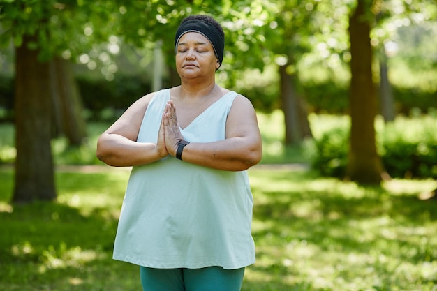 Waist up portrait of mature black woman doing yoga outdoors in green park and holding hands together