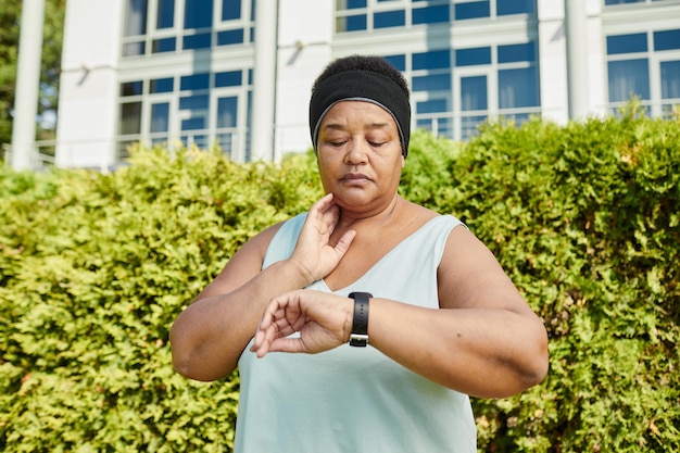 Waist up portrait of mature black woman looking at smartwatch and tracing pulse while taking break i