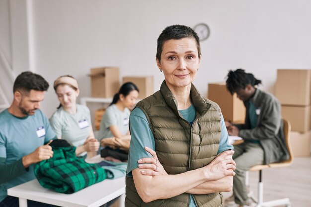 Waist up portrait of mature woman looking at camera during help and donations event with diverse team of volunteers in background, copy space