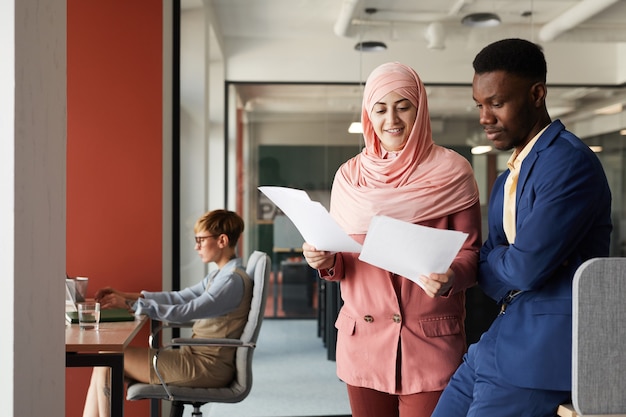 Waist up portrait of modern Muslim businesswoman talking to African-American colleague and discussing documents while standing in office interior , copy space