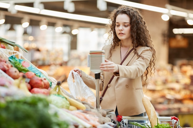 Waist up portrait of modern young woman using electric scales to weigh fresh fruits while buying groceries at supermarket