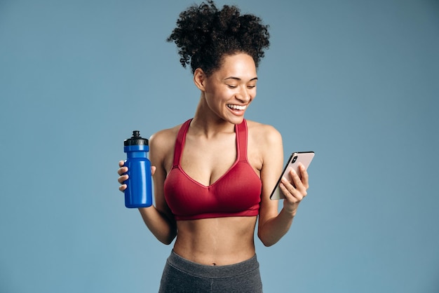 Waist up portrait of the multiracial woman holding bottle of water and checking time at her smartphone while standing in front of the camera