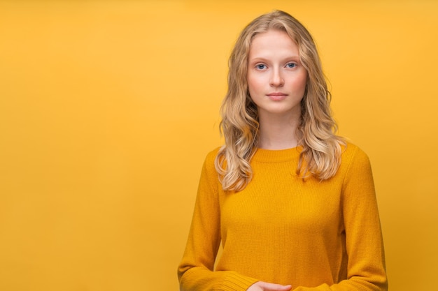Фото Талия вверх по портрету красивой молодой серьезной умной блондинки скандинавской женщины на ярко-желтой оранжевой стене. свежее лицо девушки без макияжа, вьющиеся волосы, желтый джемпер, подросток в оранжевом свитере
