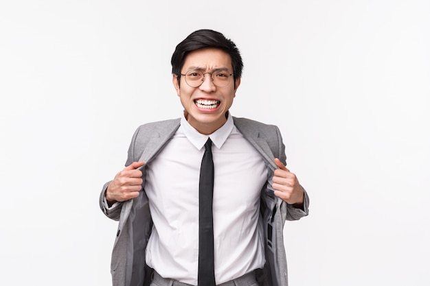 Waist-up portrait of pissed-off, aggressive young asian businessman in grey suit and tie, ripping his clothes from anger, losing patience, distressed standing over  on white wall