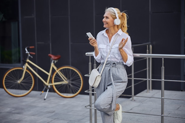 Waist up portrait of pretty lady talking with friends on mobile phone while she is standing in front of modern building and retro bike