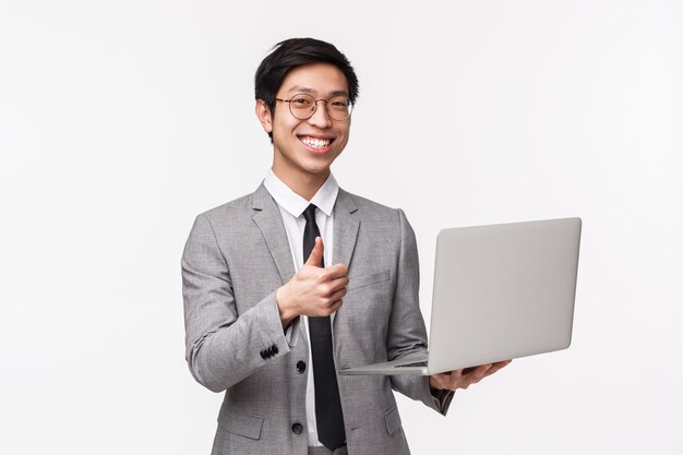 Photo waist-up portrait of satisfied, happy and pleased young asian male entrepreneur, it programmer in suit, introduce his new project, show thumbs-up in approval and holding laptop, on a white wall