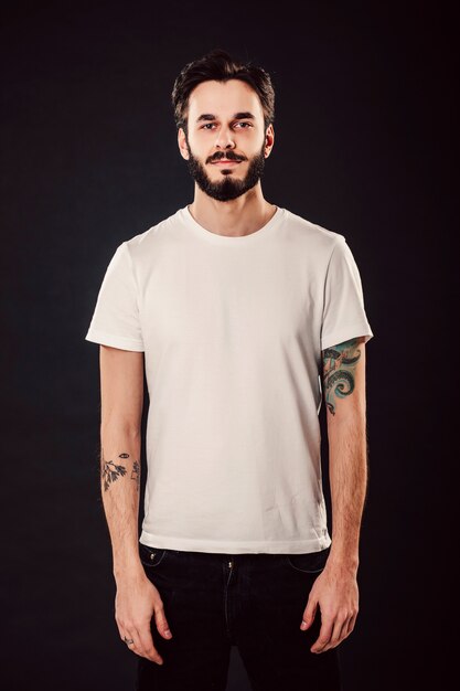 Waist up portrait of serious stylish attractive man with thick beard looks confidently, dressed in casual white t shirt