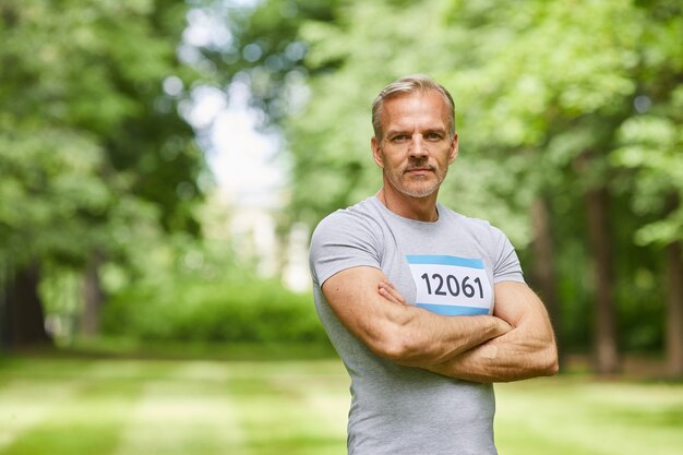 Waist up portrait shot of confident Caucasian senior sportsman taking part in summer marathon standing in park with arms crossed, copy space