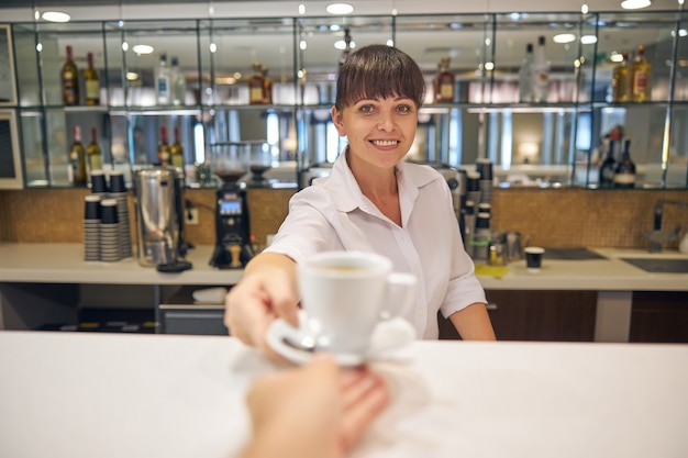 バーで働いている笑顔の美しい女性と男性に一杯のコーヒーを与える肖像画を腰に当てる