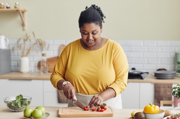 Waist up portrait of smiling black woman cooking healthy meal\
in kitchen and cutting vegetables copy