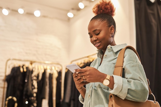 Waist up portrait of smiling black woman using smartphone in clothing store and enjoying shopping