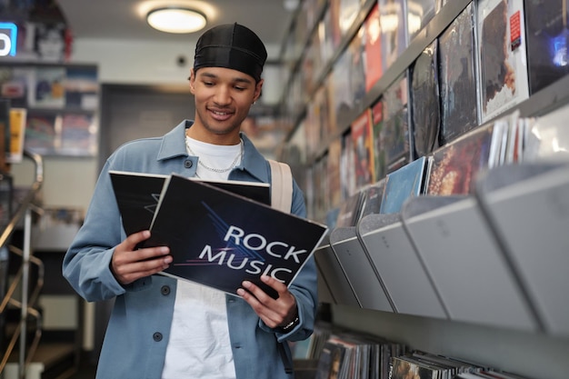 Поясной портрет улыбающегося темнокожего молодого человека, выбирающего виниловые пластинки в копировальном пространстве музыкального магазина