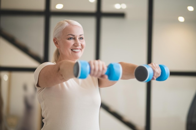 Waist-up portrait of a smiling cheerful beautiful mature blonde female doing a dumbbell front raise