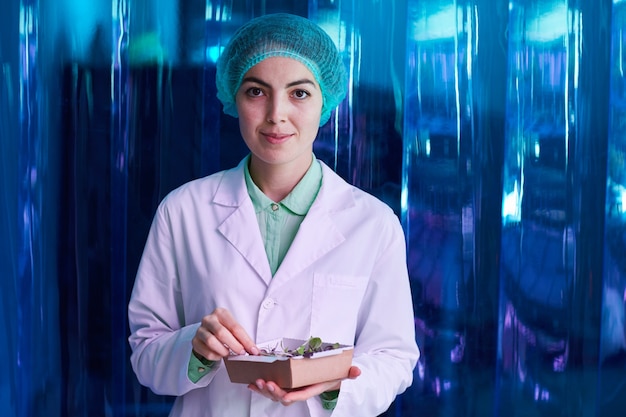 Waist up portrait of smiling female scientist holding box of green plants while posing against blue plastic wall, copy space