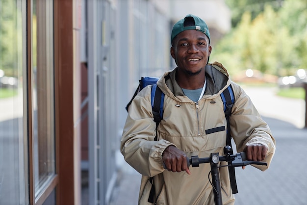 Waist up portrait of smiling food delivery worker riding electric scooter in city street