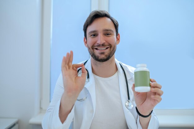 Waist-up portrait of a smiling friendly physician with the plastic vitamin bottle in his hand making the OK sign