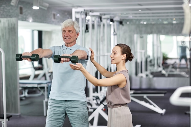 Поясной портрет улыбающегося пожилого мужчины, делающего физические упражнения в реабилитационной клинике с женщиной