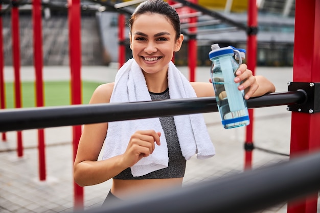 Waist up portrait of smiling sporty female with bottle of water standing on boxer ring in open air