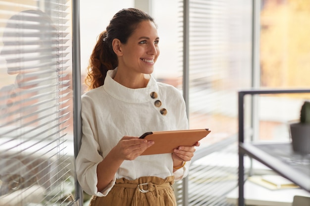 Waist up portrait of smiling successful businesswoman holding digital tablet and looking away while working in office, copy space