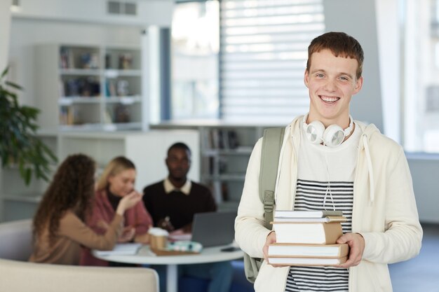 Mezzo busto ritratto di sorridente ragazzo adolescente mentre si sta in piedi nella biblioteca del college con persone che lavorano in background,