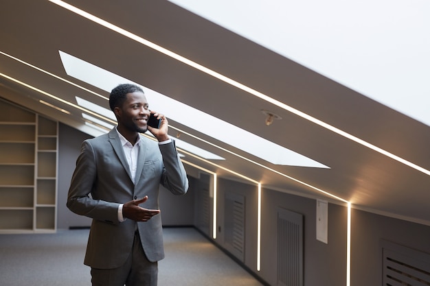 Waist up portrait of successful African-American businessman speaking by smartphone while standing in minimal grey interior of modern office lit by skylights, copy space