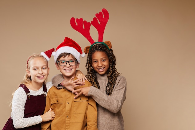 Mezzo busto ritratto di tre bambini che indossano cappelli di babbo natale sorridendo alla telecamera mentre in piedi su sfondo beige, copia spazio