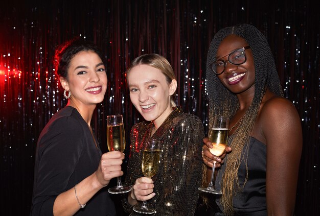 Mezzo busto ritratto di tre giovani donne eleganti che tengono bicchieri di champagne e sorridendo alla telecamera mentre posa su sfondo scintillante alla festa, girato con flash