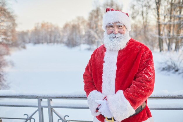 Waist up portrait of traditional santa claus looking at camera and smiling in winter park copy space