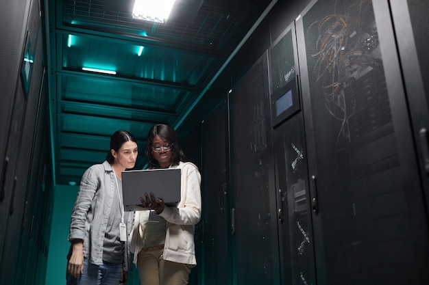 Waist up portrait of two female IT engineers setting up server network via laptop while working in data center, copy space